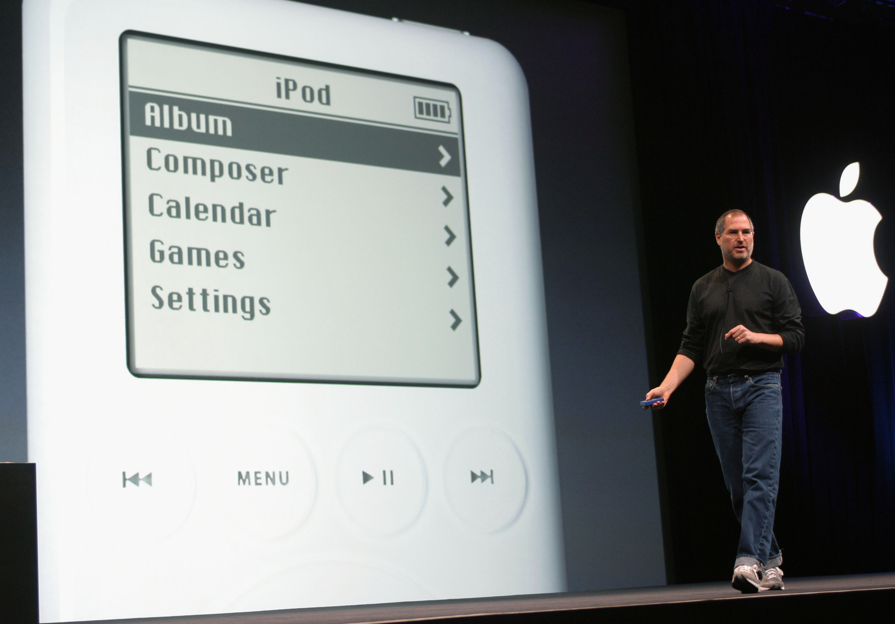 Steve Jobs on a darkened stage beside a giant projection of the original iPod’s menu screen.