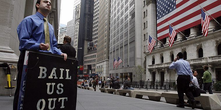 A man on Wall Street holding a “BAIL US OUT” sign beside the flag-draped New York Stock Exchange.