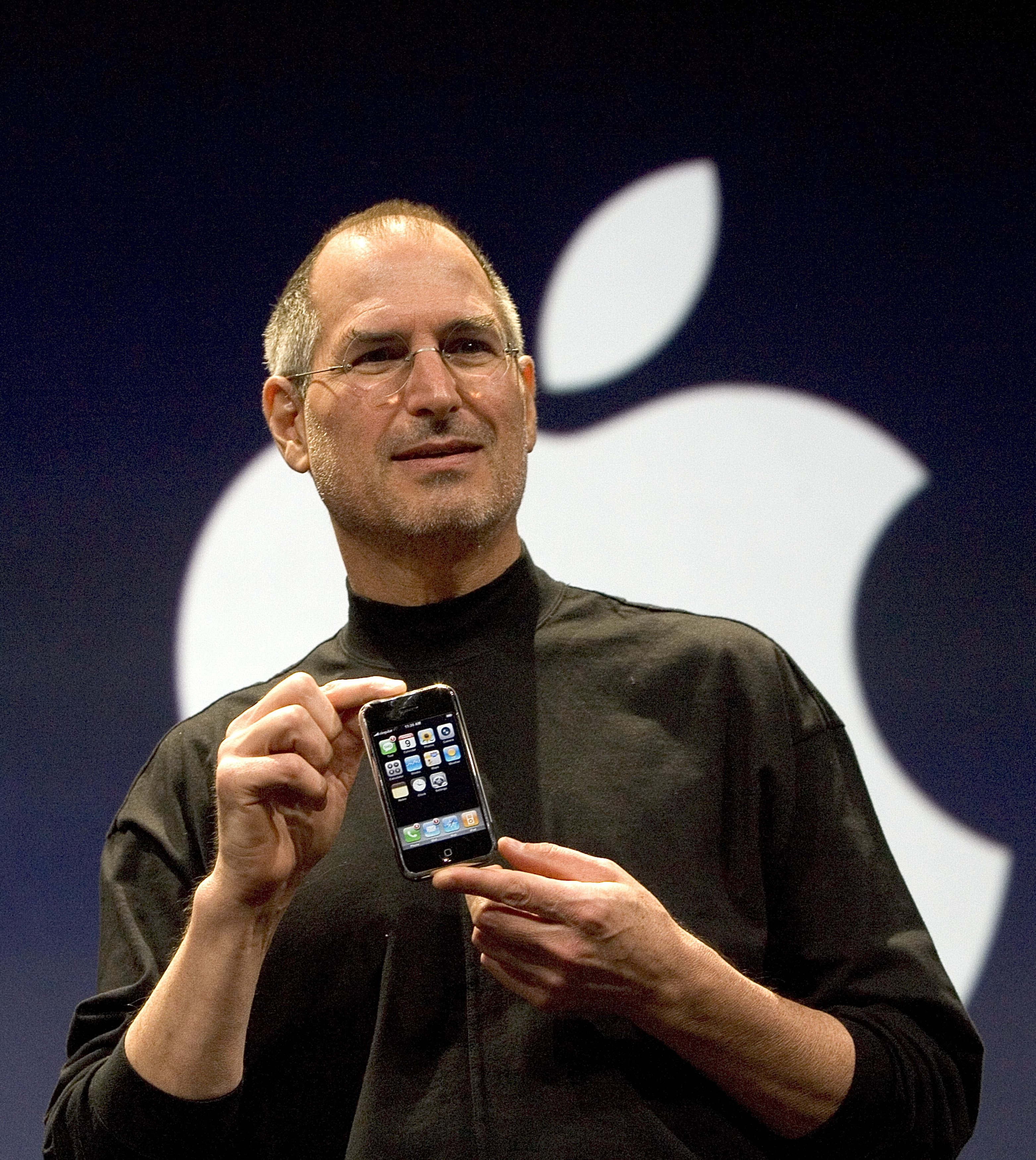 Steve Jobs holding up the first iPhone in front of a large white Apple logo.