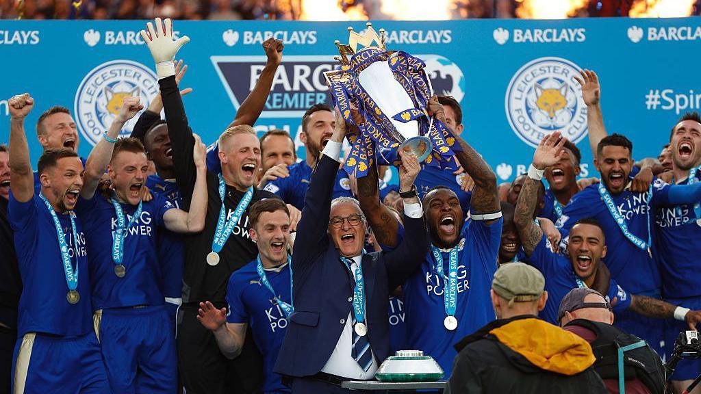 Leicester City’s players and manager Claudio Ranieri lifting the Premier League trophy amid blue confetti.