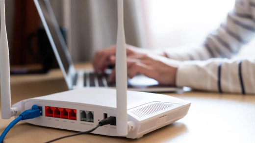 A white home broadband router with two antennae on a desk while someone types on a laptop behind it.