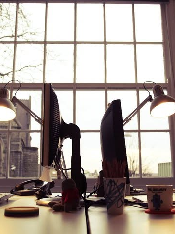 A desk silhouetted against a tall Georgian sash window, flanked by two angle-poise lamps, with flat-screen monitors and a church visible outside.