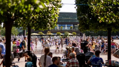 Crowds packed around a sunlit public fountain plaza on a sweltering summer day.