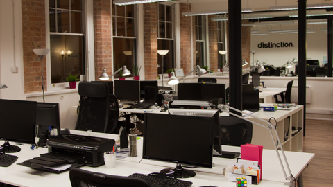 A dimly-lit open-plan office in a converted building with exposed brick, black pillars and rows of desks with monitors, “distinction.” on the far wall.