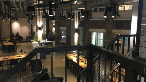 A view from a mezzanine over a dark, industrial-style open-plan office with exposed ductwork, brick and a black metal railing.