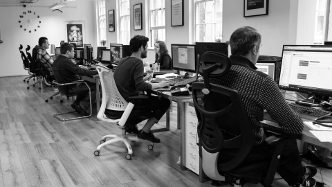 A black-and-white photo of the team working along a row of desks and monitors in a bright, window-lined office.