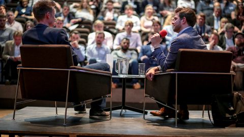 Two men in a Q&A on a stage, one holding a microphone, in front of a large seated audience.