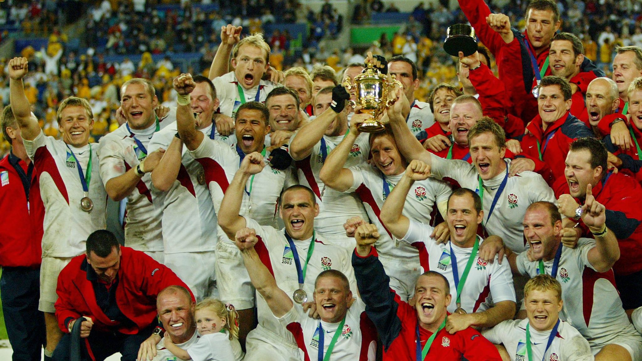 The England rugby squad celebrating on the pitch, hoisting the 2003 Rugby World Cup trophy.